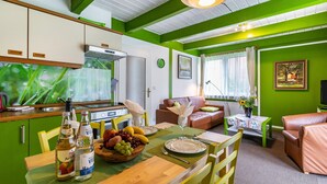 Um quarto com uma mesa de jantar posta para dois, um sofá, uma mesinha com um vaso, e uma área de cozinha com armários e um fogão.