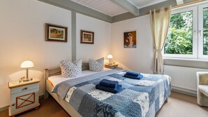 Une chambre à coucher avec un lit, des tables de chevet, des lampes, une fenêtre avec des rideaux et des tableaux encadrés accrochés au mur.