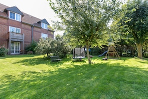 Un jardin à l’arrière d’une maison, aménagé pour les loisirs, avec un trampoline, un toboggan, une cabane en bois et une maison en briques.