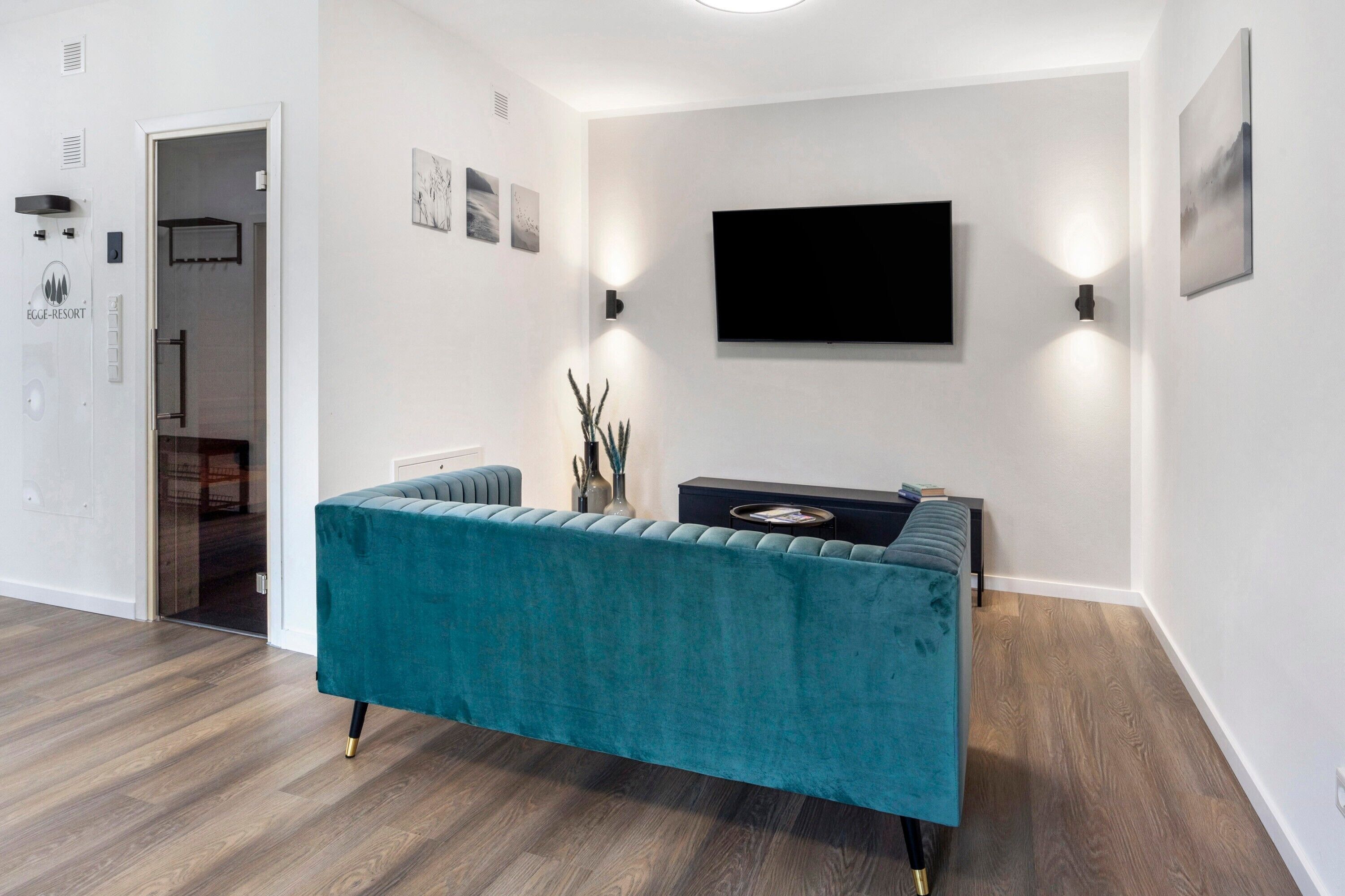 Ein modernes Wohnzimmer mit einem petrolfarbenen Sofa, einem flachbildfernseher an der Wand und einem kleinen Beistelltisch.