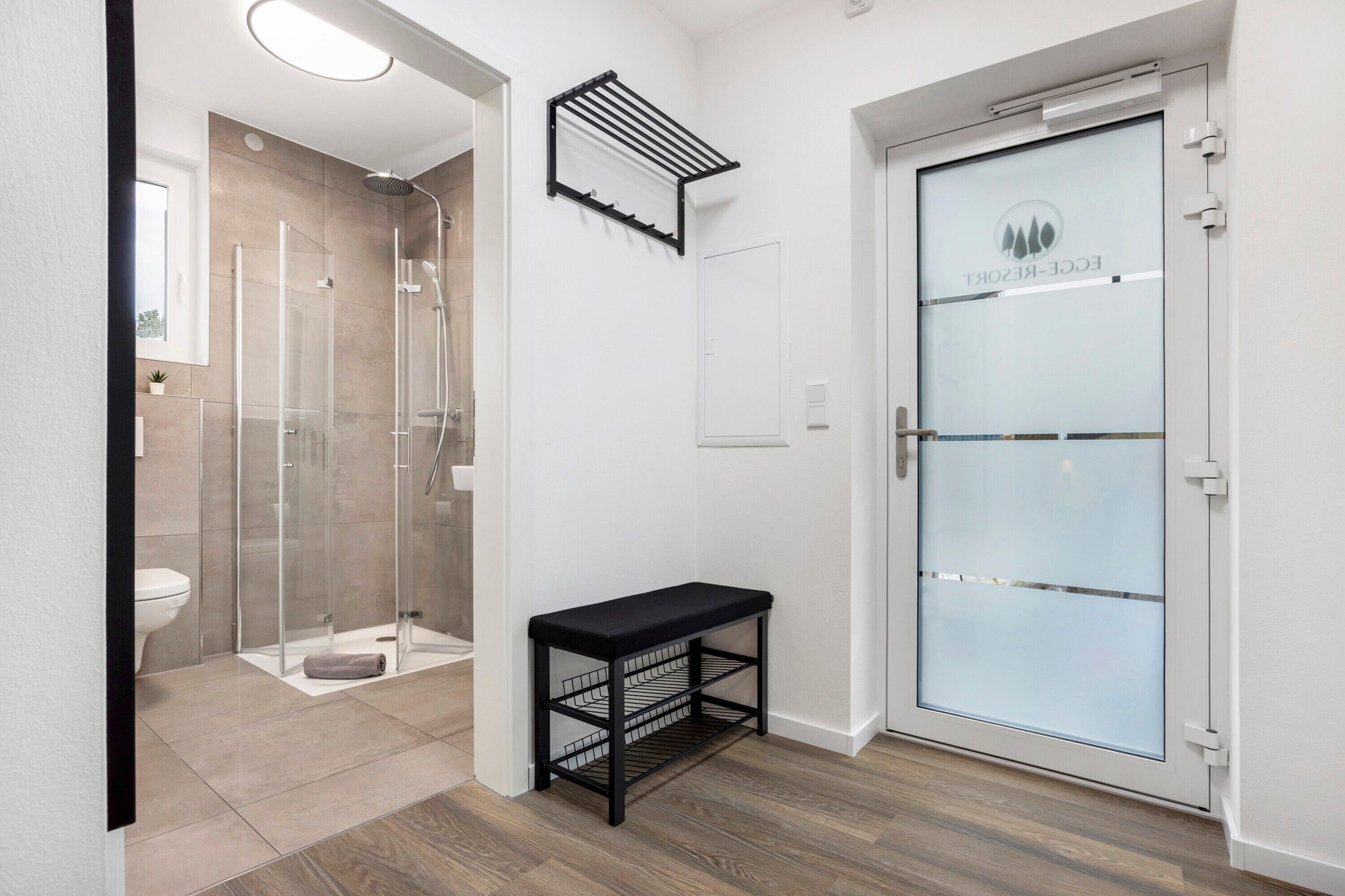 A modern bathroom with a glass shower enclosure, a bench with storage, and a frosted glass door with a logo.