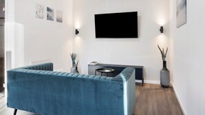 Una sala de estar moderna con un sofá azul, un televisor de pantalla plana montado en la pared, una mesita auxiliar y dos jarrones con plantas.
