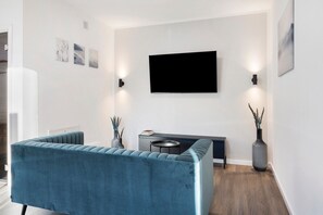 A modern living room with a blue sofa, a flat-screen TV mounted on the wall, a small side table, and two vases with plants.
