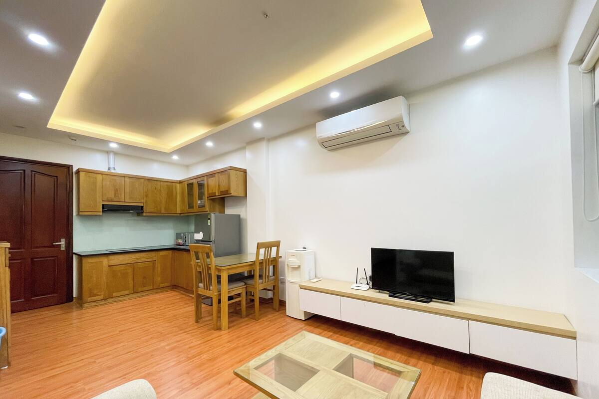 Un appartement moderne comprenant une cuisine, un coin repas et un salon. Il est équipé d’armoires en bois, d’un téléviseur à écran plat, d’une table basse en verre et d’un système de climatisation intégré.
