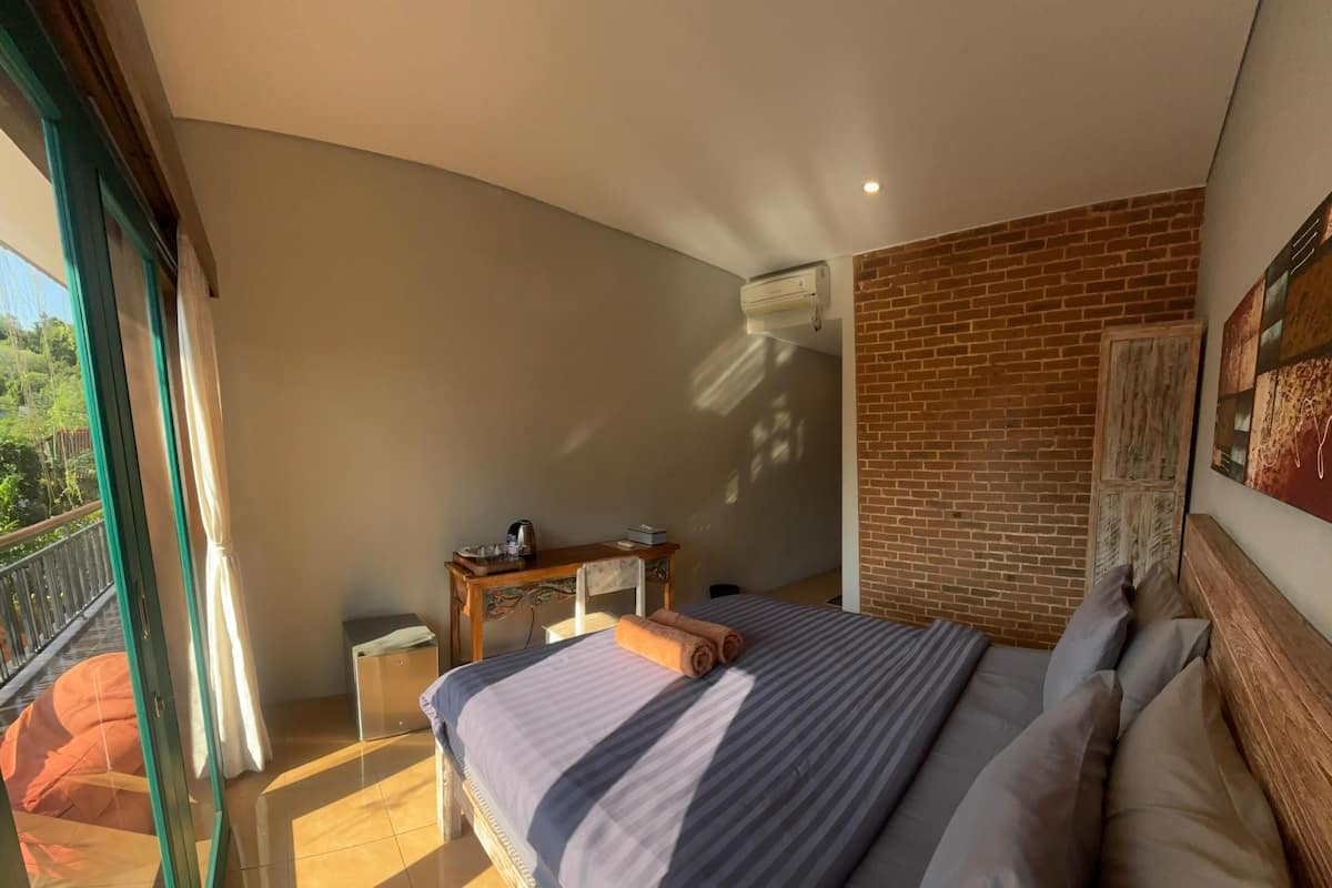 A bedroom with a bed, a table, and a chair. There is a balcony with a view of greenery.