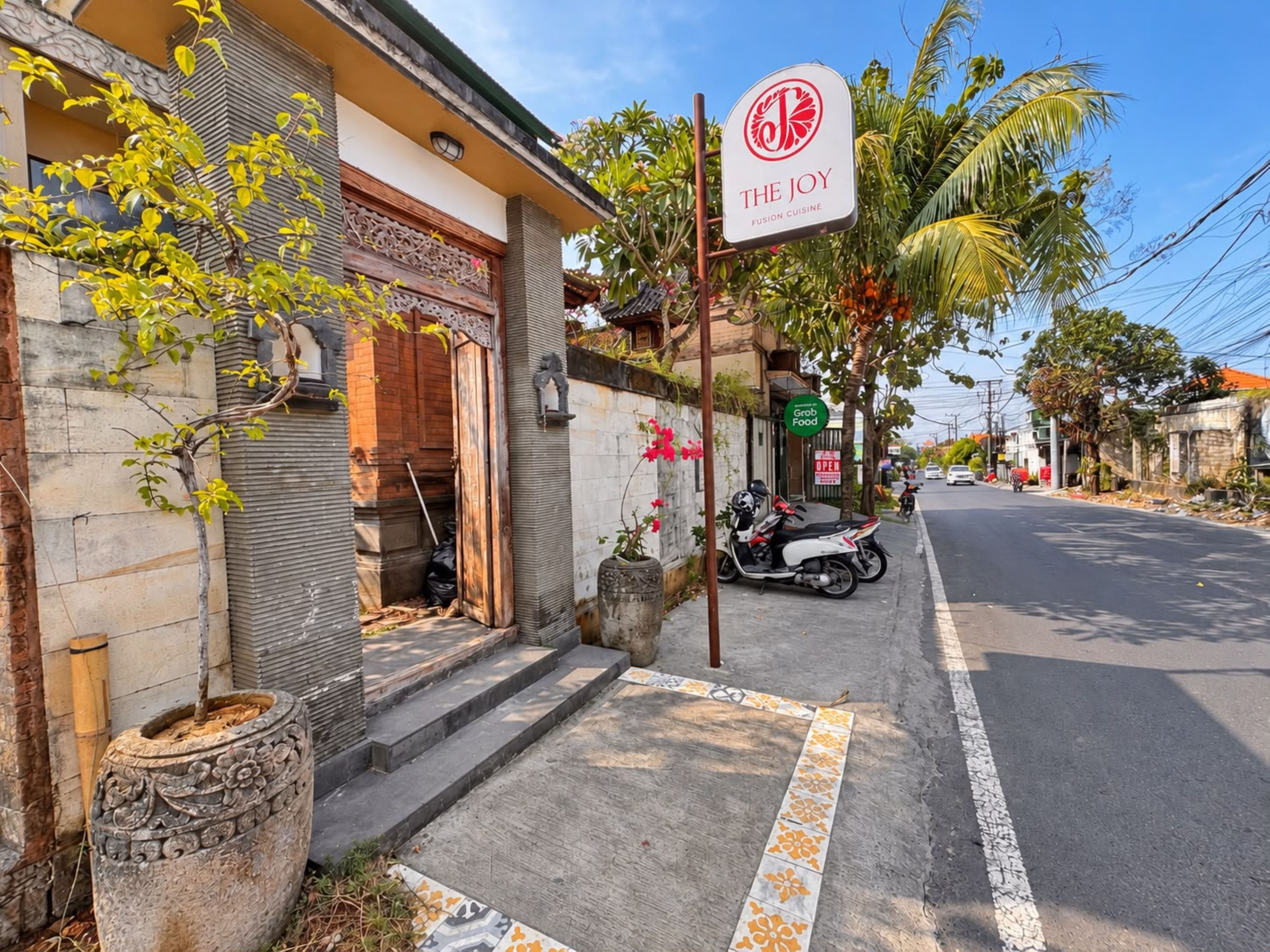A street view with a restaurant named 'The Joy Fusion Cuisine' sign, a scooter parked on the sidewalk, and a street lined with palm trees and residential buildings.