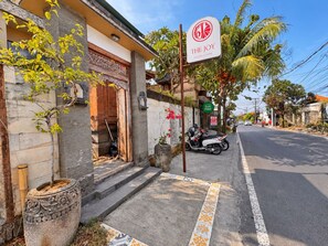 Straßenansicht mit einem Restaurant namens 'The Joy Fusion Cuisine', einem auf dem Bürgersteig abgestellten Roller sowie einer von Palmen und Wohnhäusern gesäumten Straße.