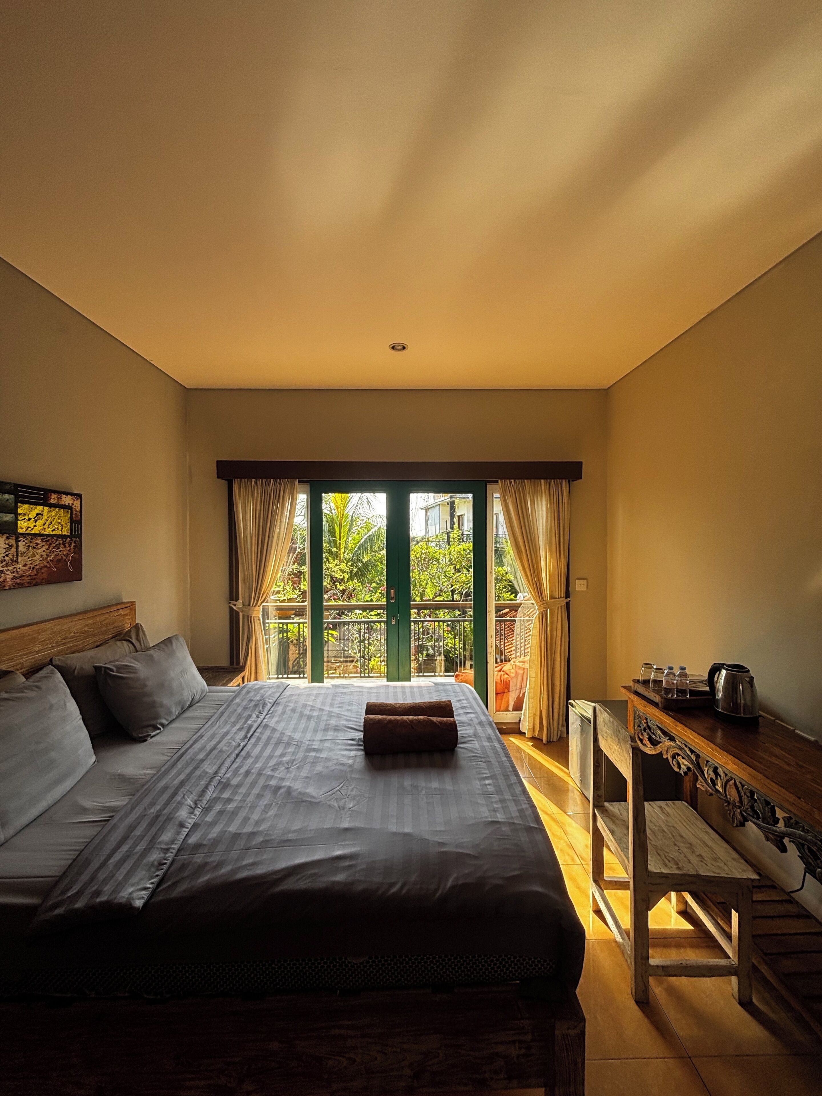 A bedroom with a large bed, a wooden nightstand, a window with curtains, and a view of greenery outside.