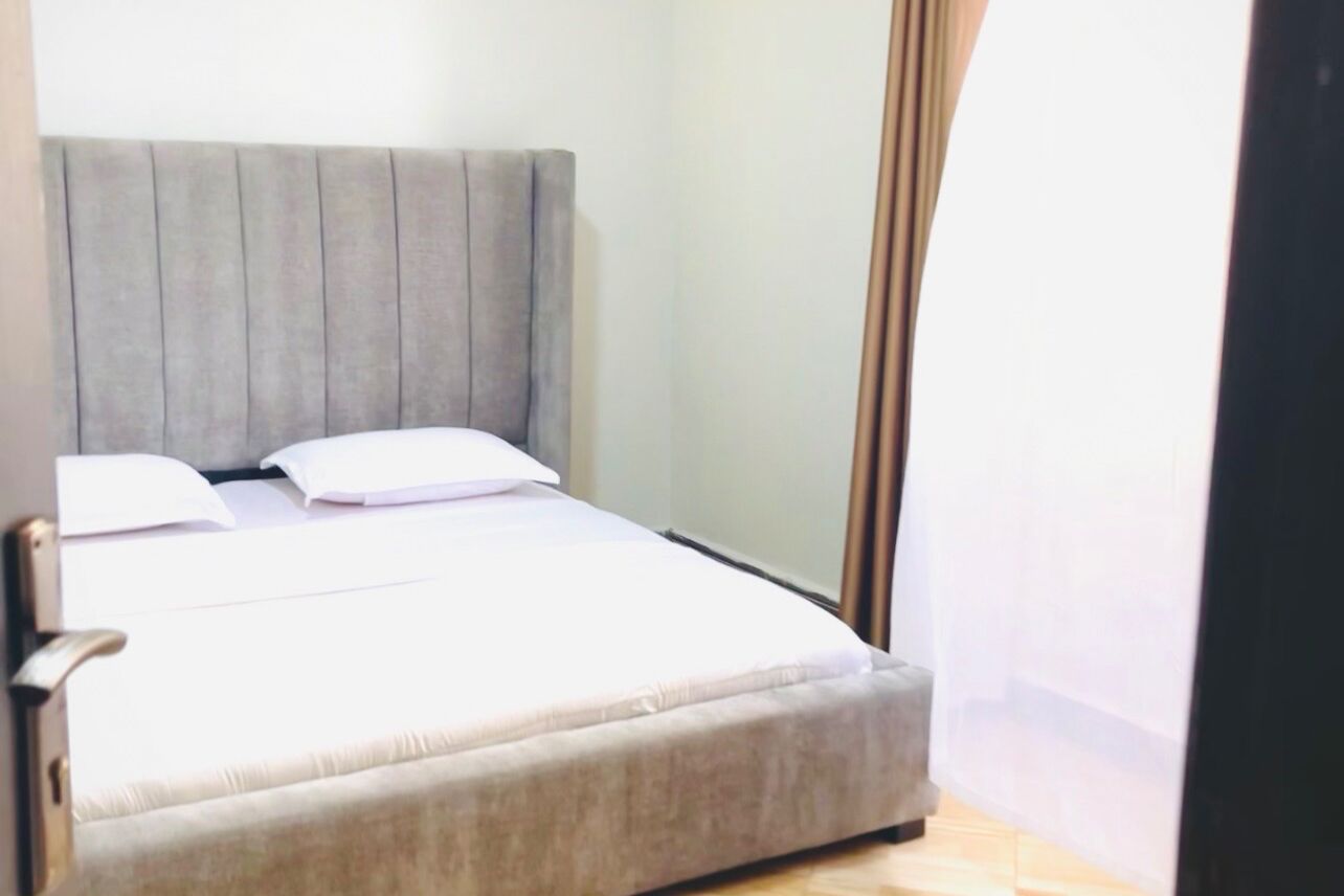 A neatly made bed with a grey headboard in a room with a plain wall and a curtain on one side.