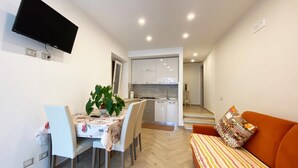 A modern living room with a sofa, a dining table with chairs, a TV mounted on the wall, and a kitchen area with cabinets and a countertop. - 1 Bed House - Sleeps 3 - Wifi (Minori)