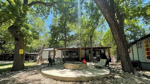 A rustic outdoor seating area with a stone fire pit, surrounded by trees and a small building with a sign.