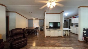 A living room with a leather sofa, a dining area with a table and chairs, a TV on a stand, and a kitchen area with cabinets and appliances.