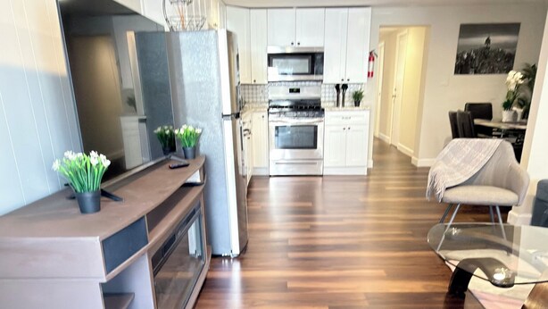 A modern kitchen with white cabinets, a microwave, and an oven. There is a dining area with a glass table and chairs, and a living room with a sofa and a coffee table. - Apartment with excellent location, a few minutes from Manhattan New York (Jersey City)