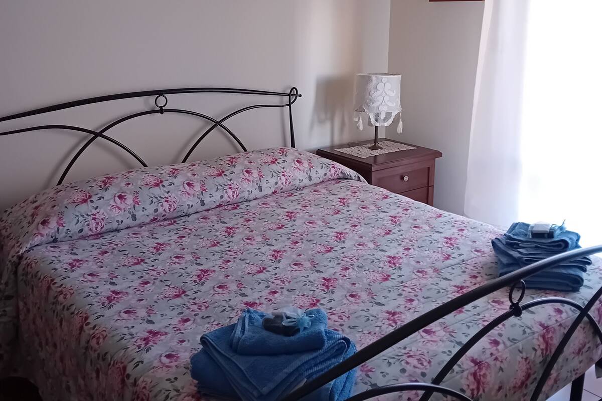 Een slaapkamer met een bed, nachtkastje, lamp en ingelijste foto’s aan de muur.