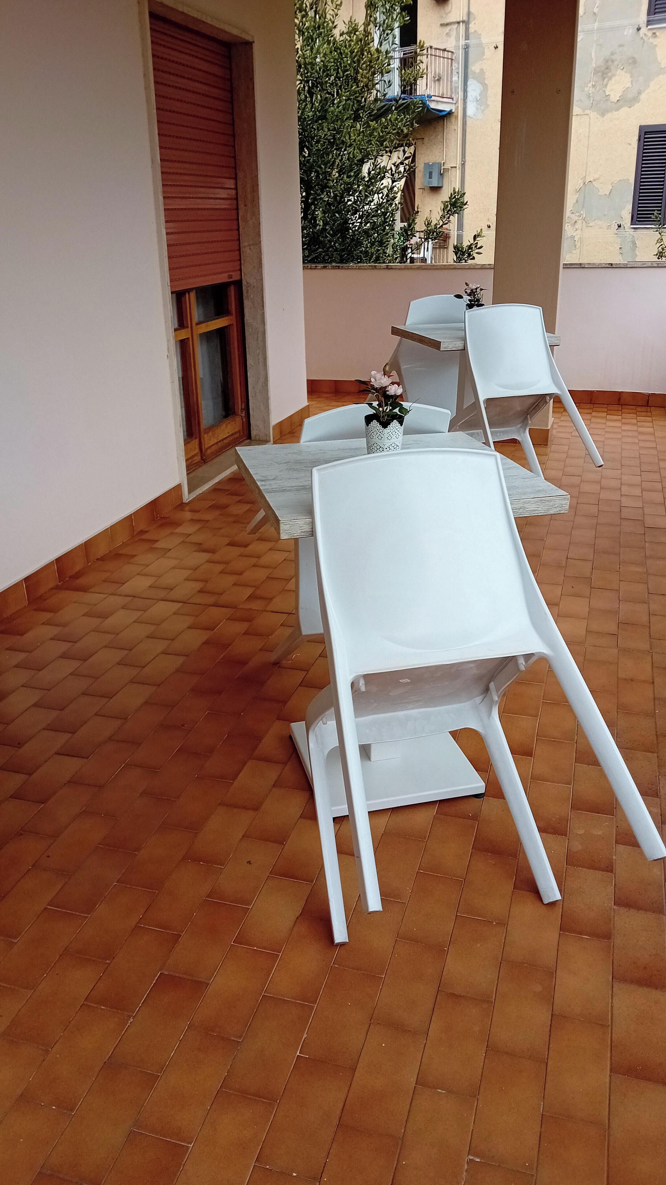 Eine Terrasse mit weißen Tischen und Stühlen, einer roten Rolltür und einem gefliesten Boden.