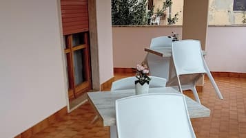 Una terraza con mesas y sillas blancas, una puerta con contraventanas rojas y suelo de baldosas.
