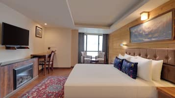 A hotel room with a large bed, a flat-screen TV, a fireplace, a dining table with chairs, and a large window.