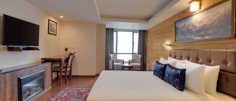 A hotel room with a large bed, a flat-screen TV, a fireplace, a dining table with chairs, and a large window.