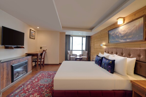 A hotel room with a large bed, a flat-screen TV, a fireplace, a dining table with chairs, and a large window.