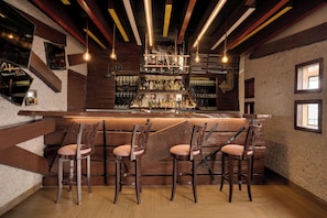 A bar area with wooden bar stools, a bar counter stocked with bottles, and a rustic ceiling with exposed beams and hanging lights.
