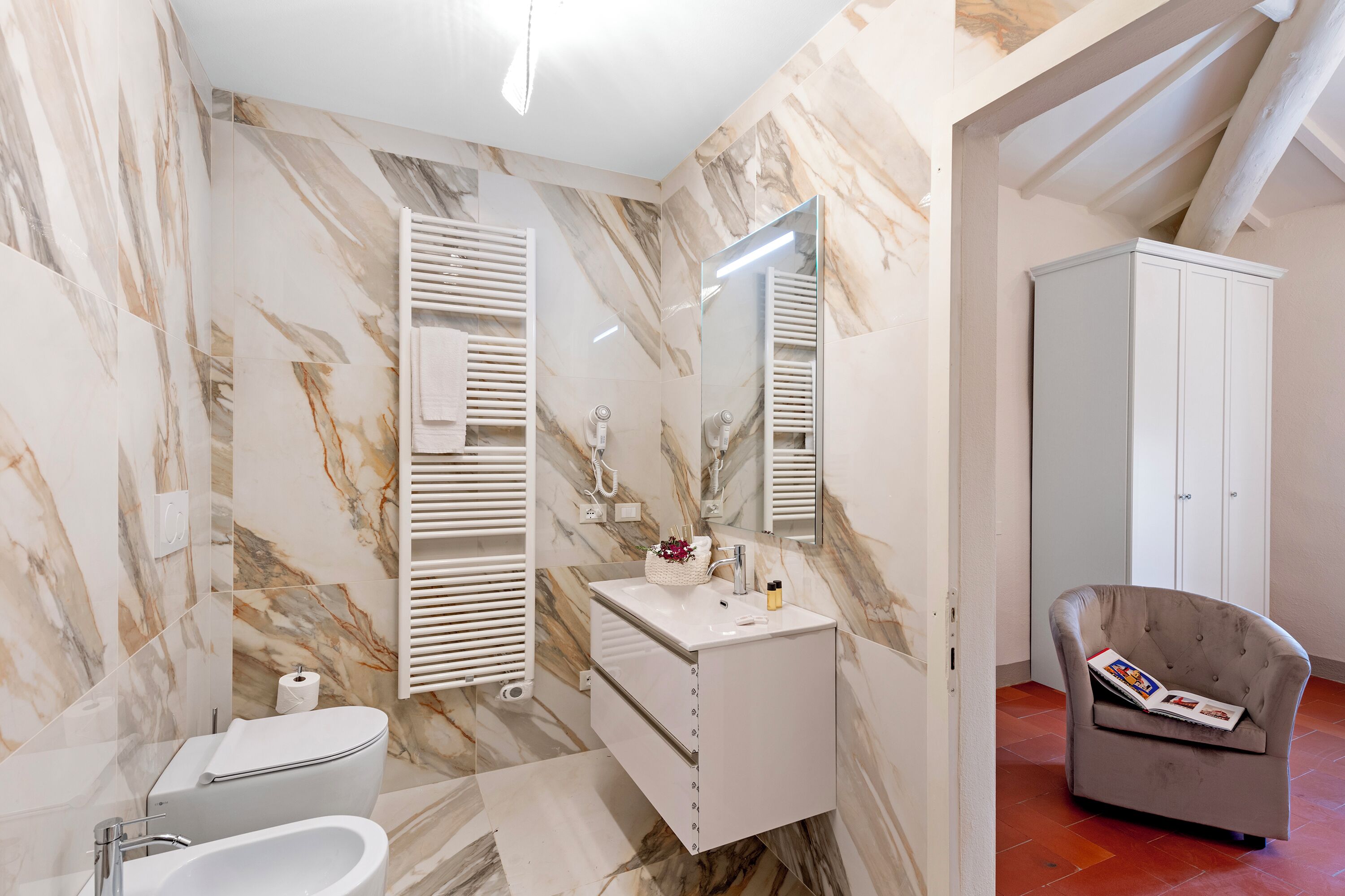 Ein Badezimmer mit Marmorwand, einem weißen Waschbecken, einer Toilette, einem Handtuchhalter, einem Schrank und einem Lesesessel.
