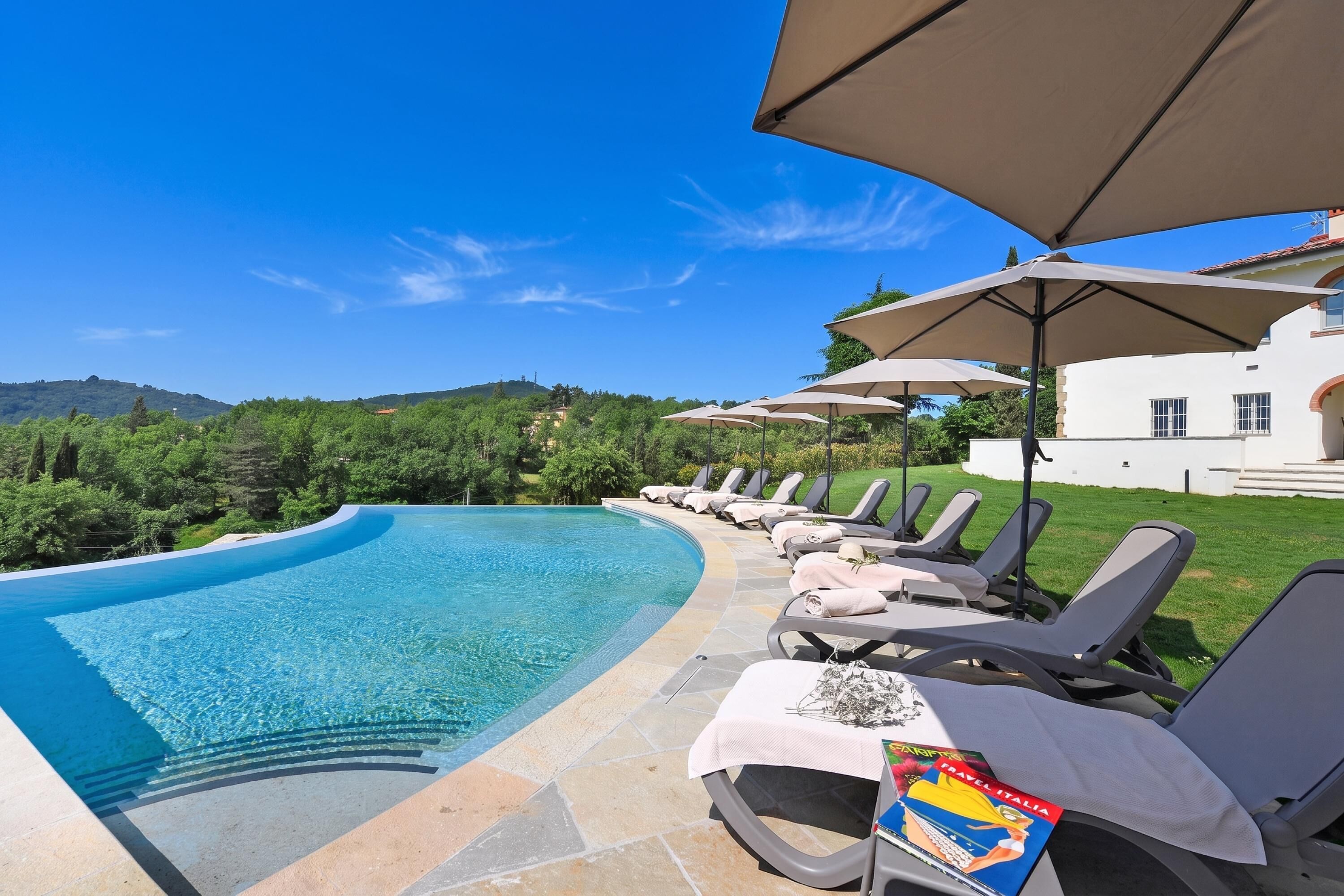 A swimming pool with lounge chairs and umbrellas, surrounded by greenery and a building.