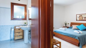 Ein Schlafzimmer mit einem hölzernen Bett, einem Nachttisch, einem Fenster und einem Badezimmer mit Waschbecken und Korbwäsche.
