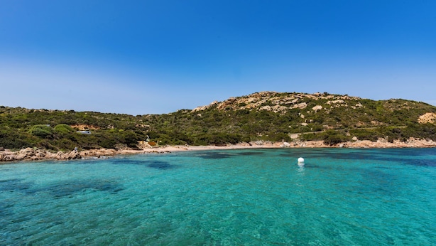 Eine Küstenlandschaft mit klarem türkisblauem Wasser, einem Sandstrand und einem Hügel mit spärlicher Vegetation.