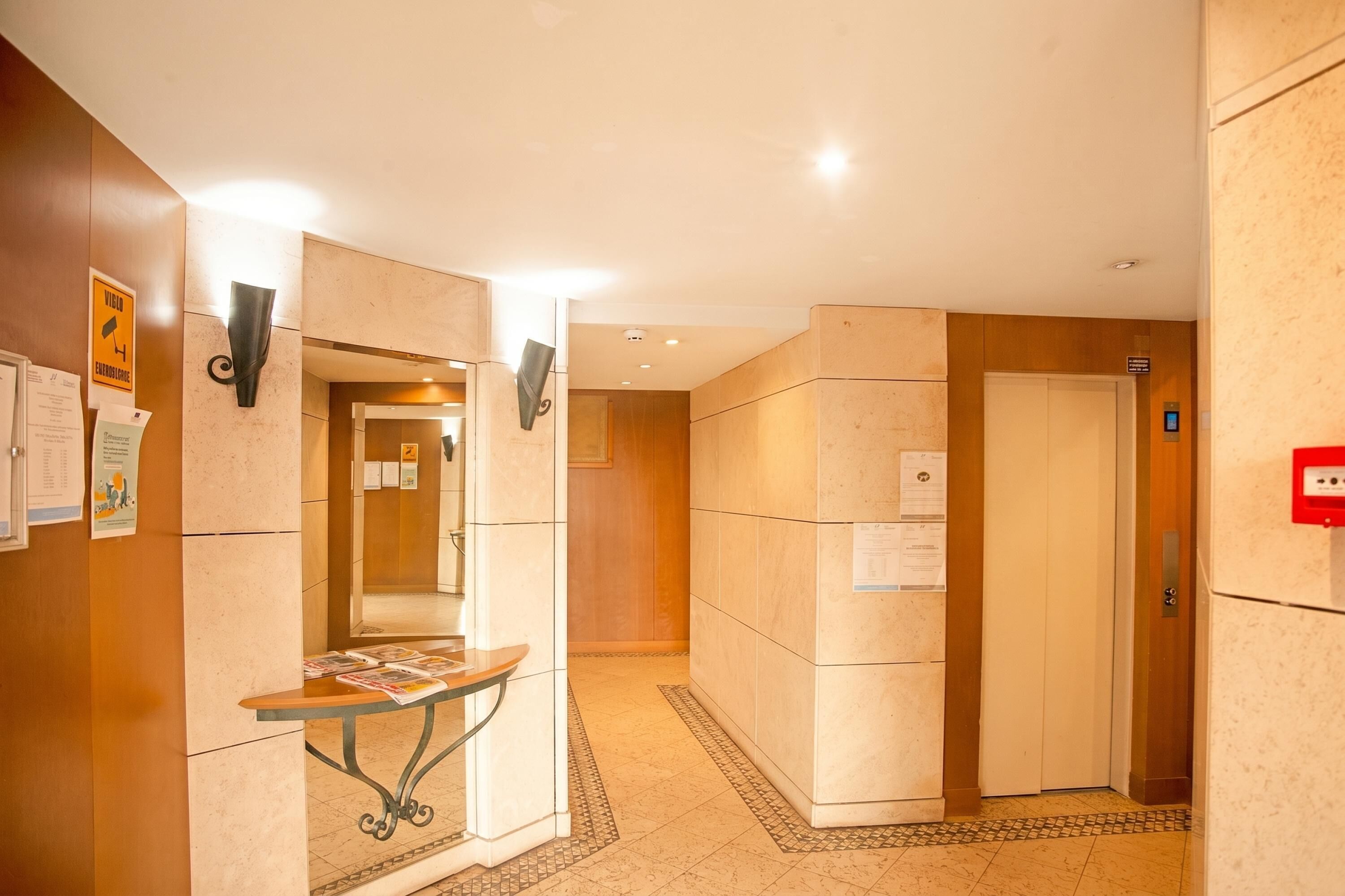 Een hotel lobby met een receptiebalie, een informatiebord aan de muur, een lift en een betegelde vloer.