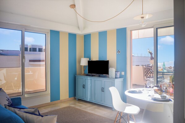 A room with a sliding glass door to a balcony, a blue and beige striped wall, a flat-screen TV on a cabinet, a dining table with chairs, and a small table with a vase and bottle.