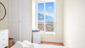 Ein Schlafzimmer mit einem Bett, einer weißen Kommode, einem Mülleimer und einem Fenster mit Blick auf das Meer und die Berge.