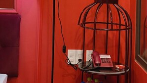 A room with a red wall, a green floor lamp, a metal birdcage with a telephone, and a small table with a bottle. - Thuong Hai Hotel - by Bay Luxury (Hanoi)