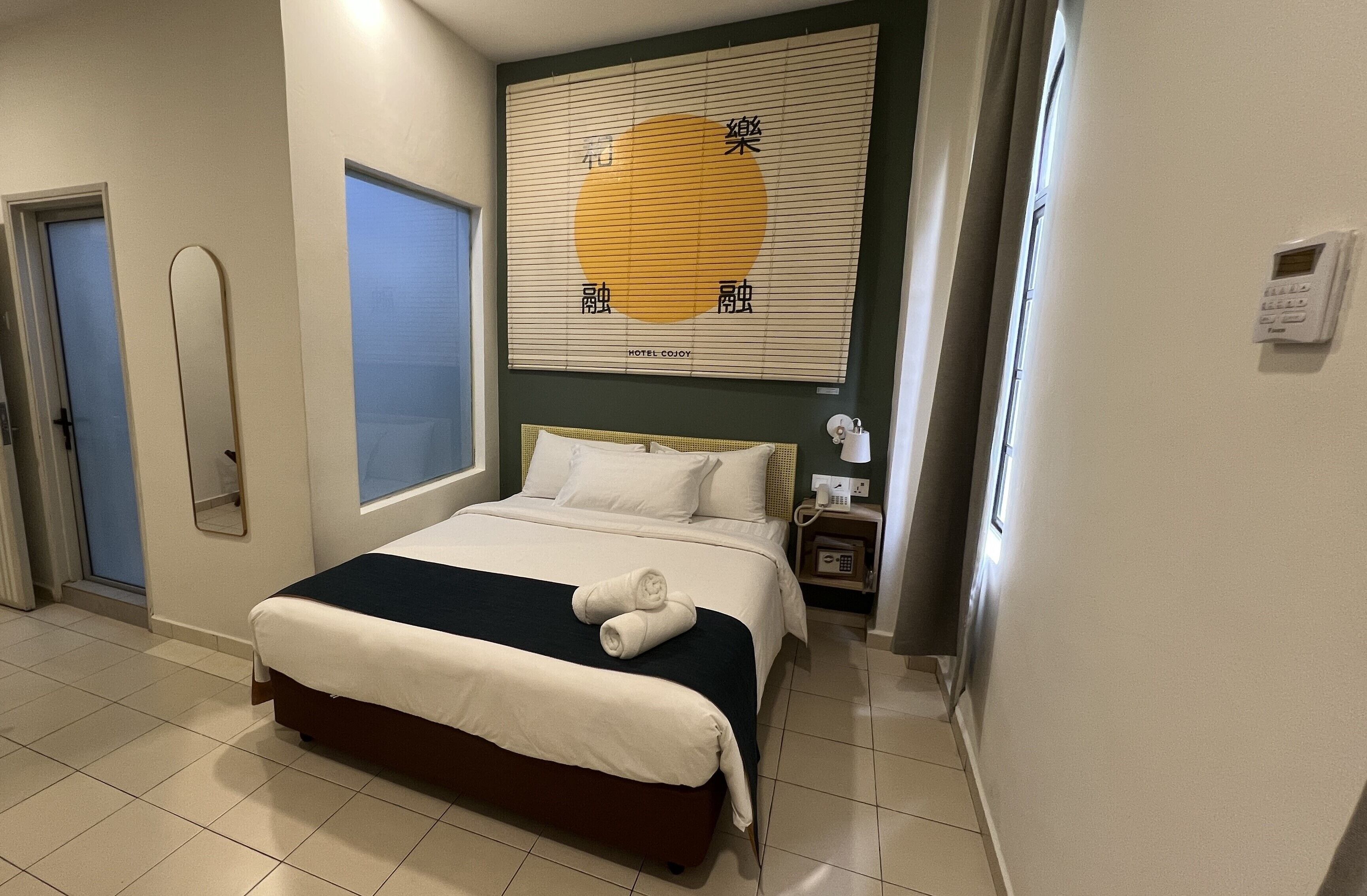 Ein Hotelzimmer mit einem Bett, einem Nachttisch mit Telefon, einem Fenster und einer Wand mit einem großen Plakat.