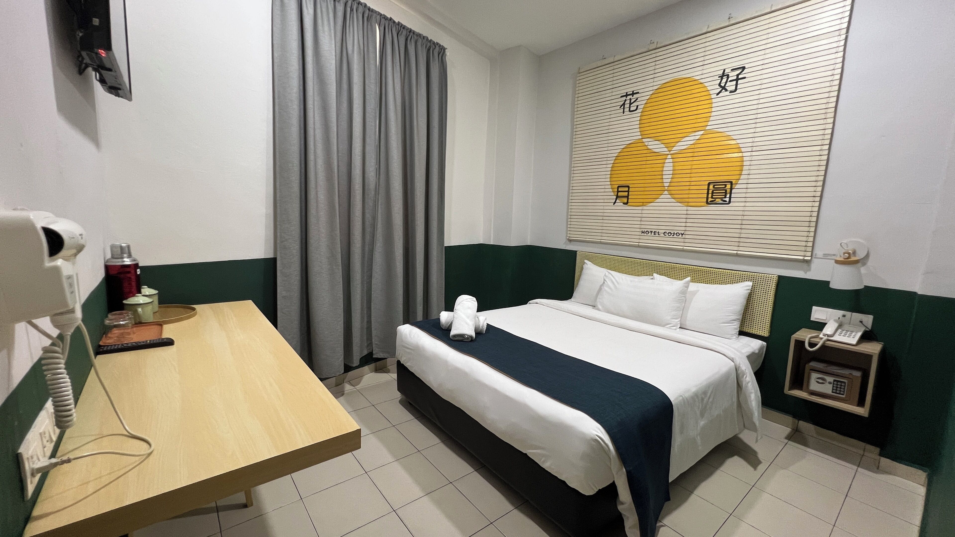 A hotel room with a bed, a table with a hairdryer, a phone, and a wall-mounted artwork.