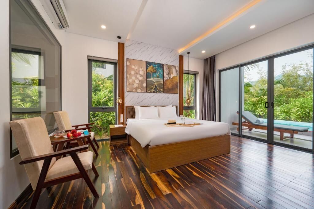 A modern bedroom with a large bed, wooden flooring, a dining table with chairs, and a view of a lush garden through large windows.