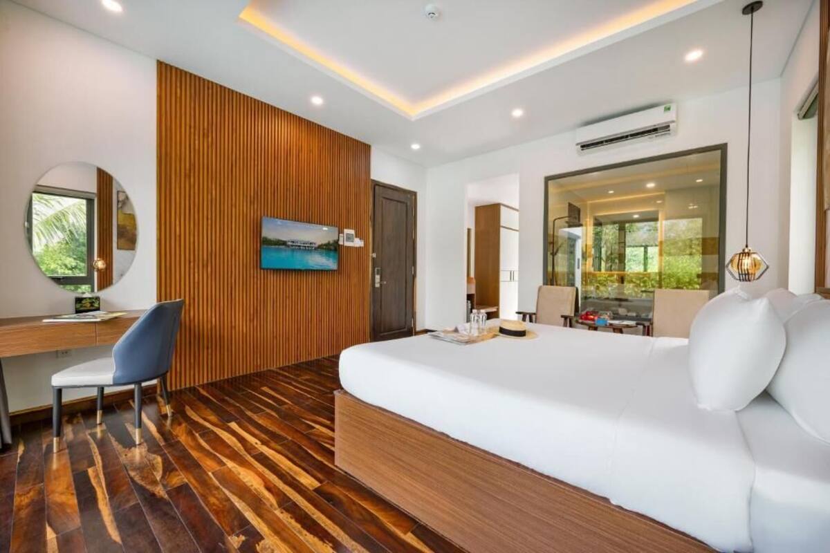 A modern hotel room with a wooden floor, a large bed, a desk with a chair, a TV, and a large window offering a view of greenery.