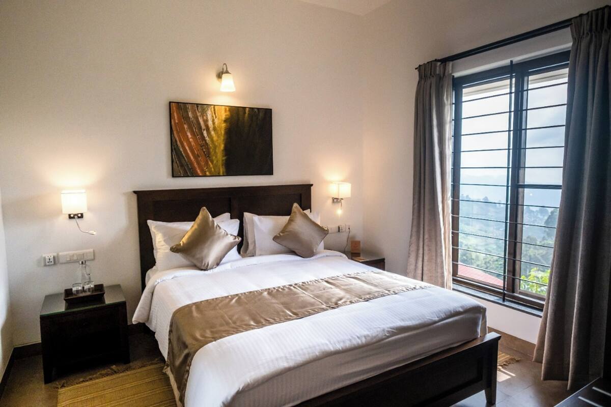 Cama bem arrumada com cabeceira, duas mesas de cabeceira com luminárias, um quadro na parede e uma janela com cortinas.