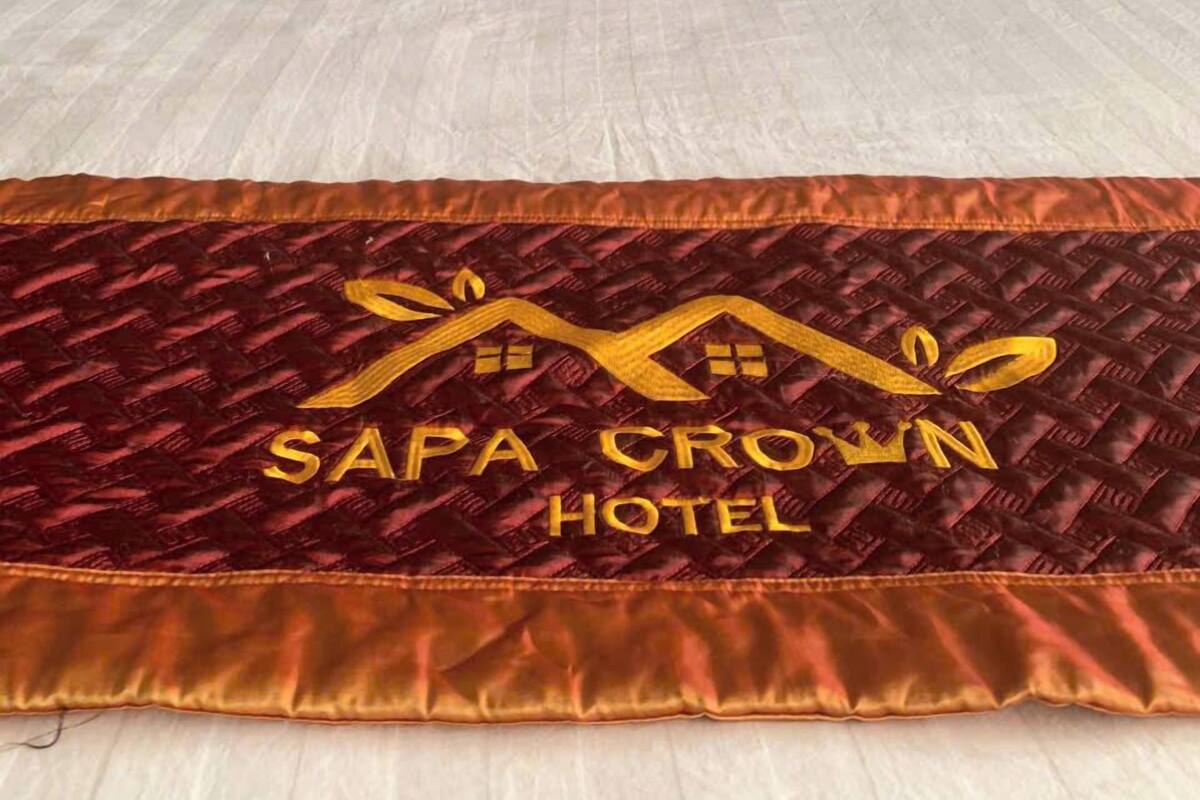 Uma cama com conjunto de almofadas branco e vermelho, uma colcha bordada dourada com o texto "SAPA CROWN HOTEL" e uma cabeceira escura.