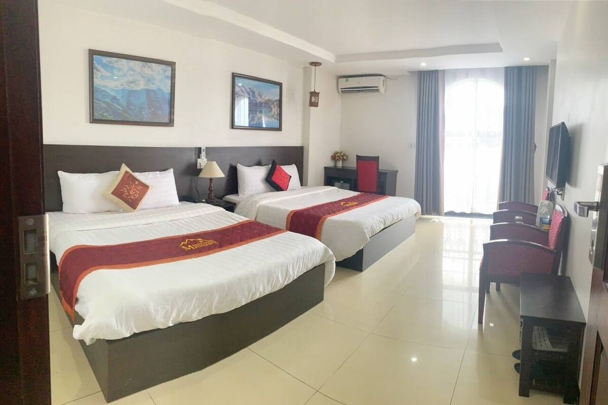 Quarto de hotel com duas camas, uma escrivaninha, uma cadeira, uma televisão e uma janela com cortinas.