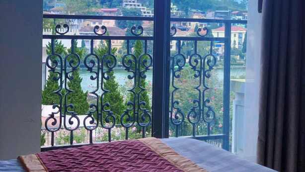 A room with a large window overlooking a balcony with decorative iron railings and a view of buildings and trees. - Sapa Crown Hotel (Sa Pa)