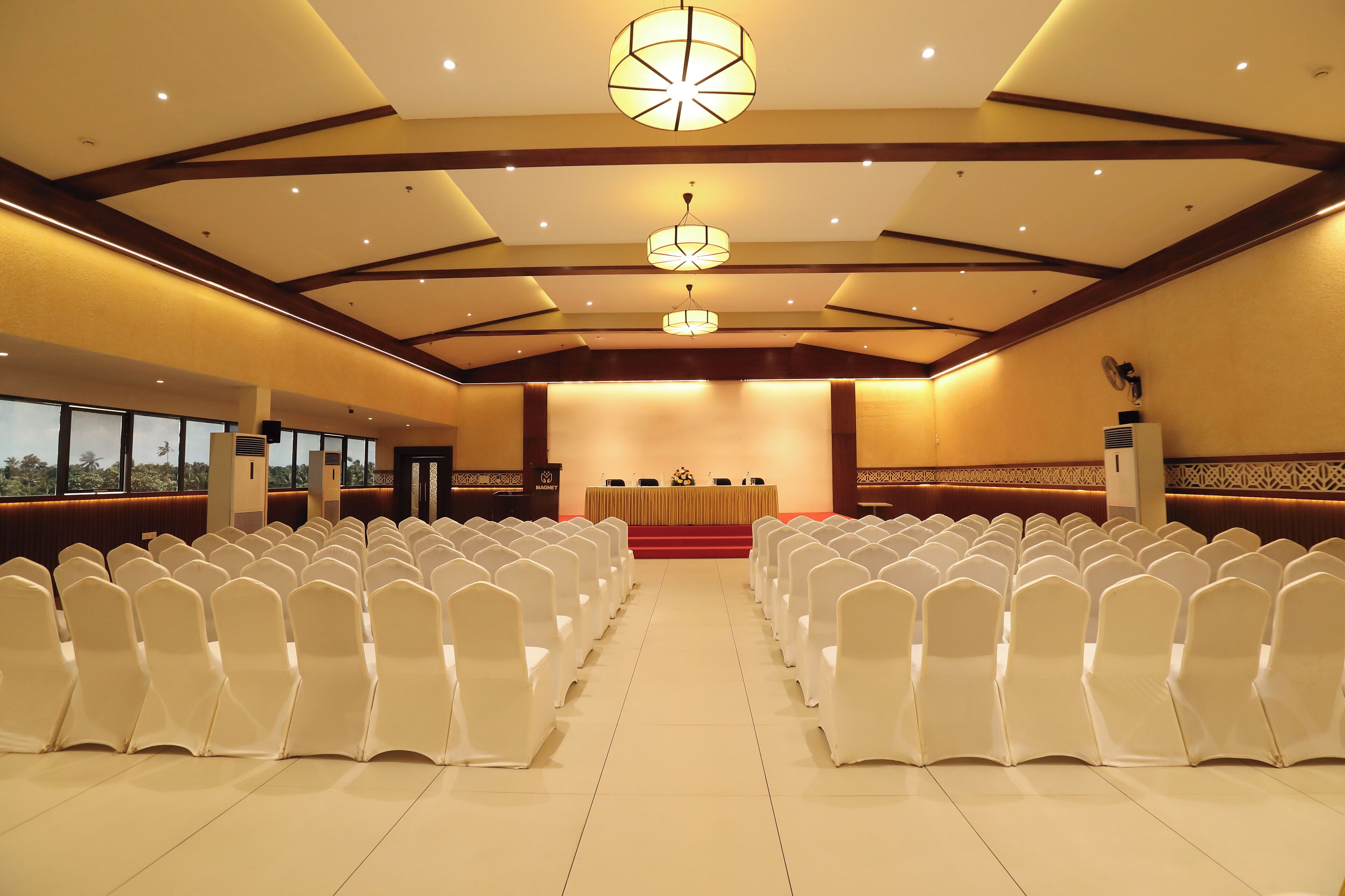 A conference room with rows of white chairs, a stage with a podium, and a large chandelier.