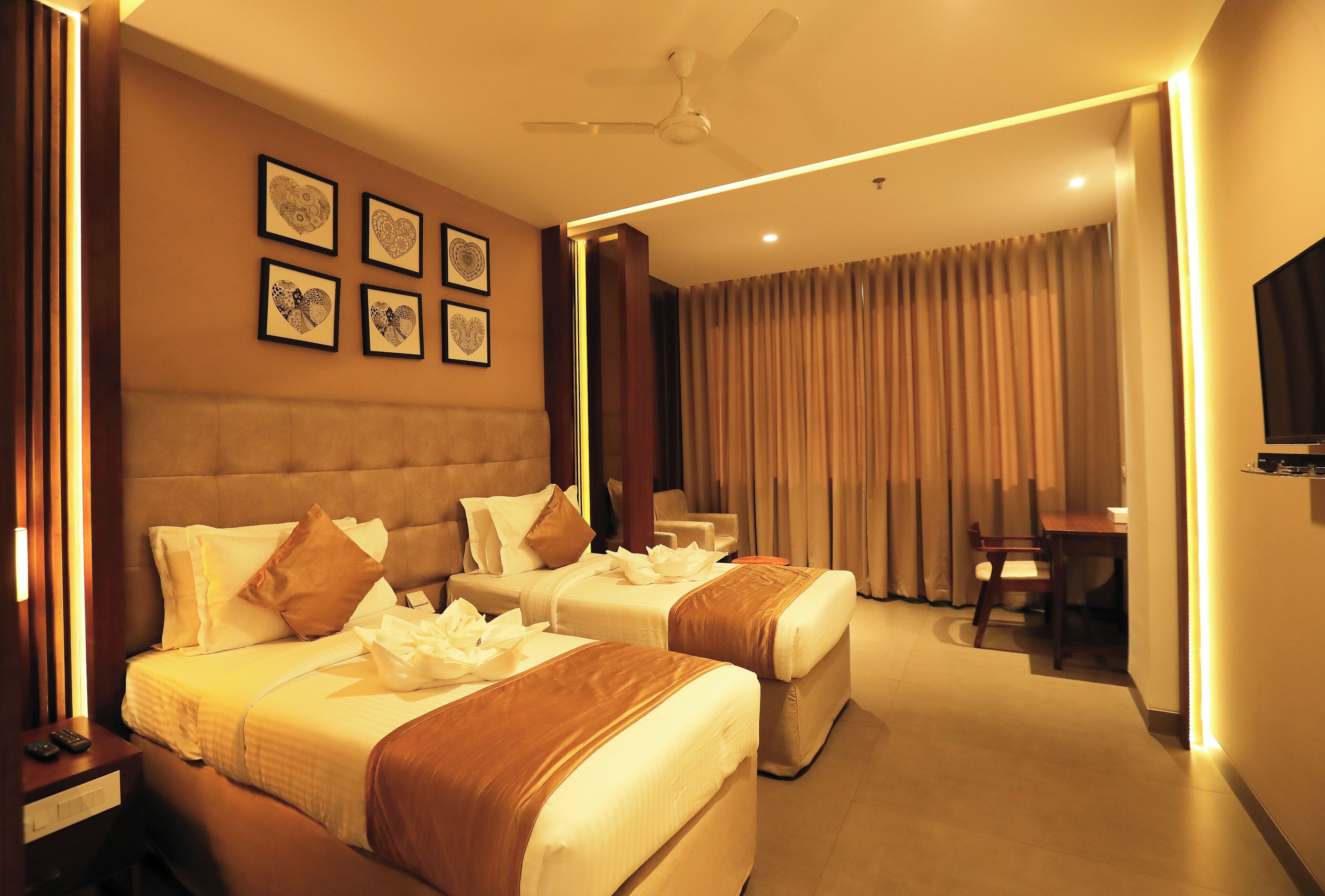A hotel room with two beds, a desk, a chair, a ceiling fan, and framed artwork on the wall.