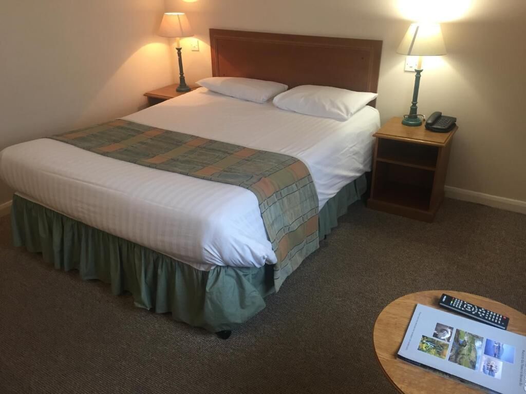 A neatly made bed with a green skirt, two bedside lamps, a nightstand with a phone, and a brochure on a round table.
