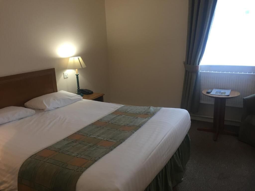 Une chambre d’hôtel avec un grand lit, une lampe de chevet, une petite table avec une chaise et une fenêtre avec des rideaux.