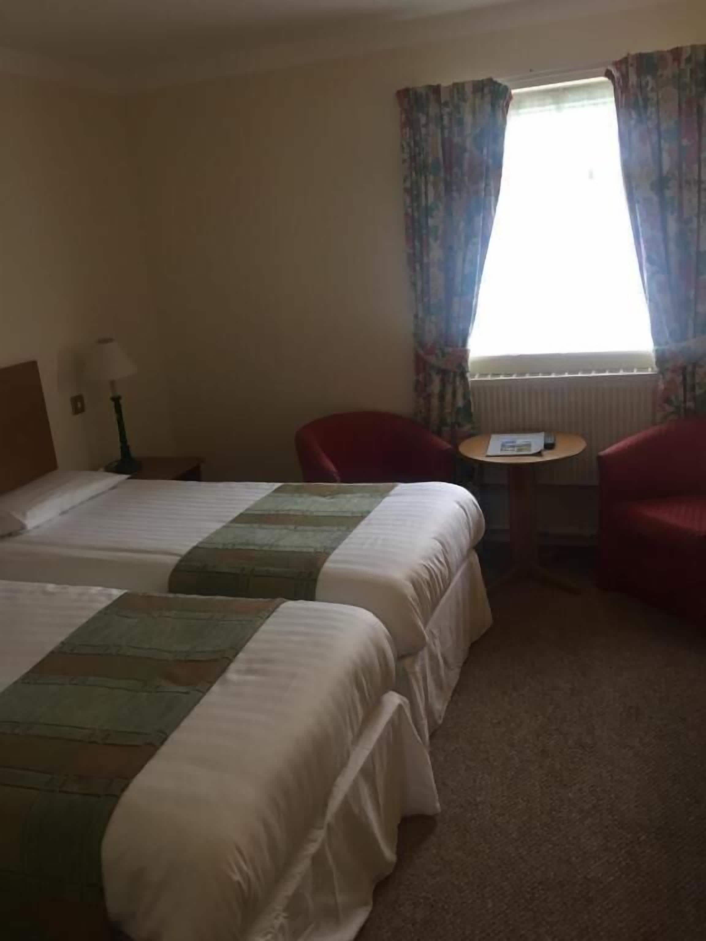 Hotellihuone, jossa on kaksi sänkyä, pieni pöytä, tuoli ja ikkuna, jossa on verhot.