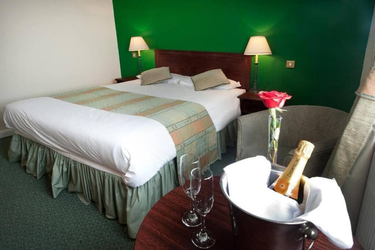 A hotel room with a bed, two bedside lamps, a small table with a vase of flowers and a bottle of champagne, and a chair.