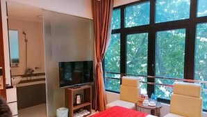 A hotel room with a bed, a TV, a small table, two chairs, a window with a view of trees, and a bathroom with a shower and sink.