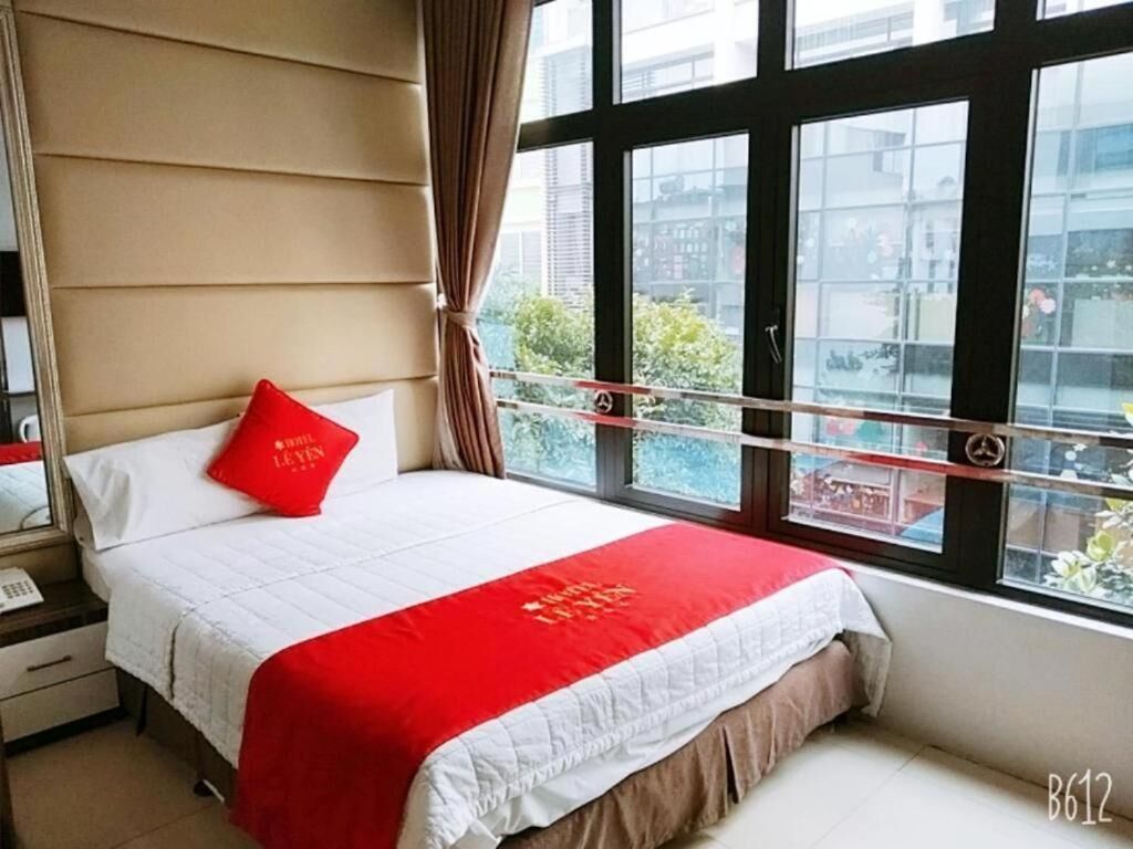 A hotel room with a bed, a red pillow, a white bedspread, and a window with a view of buildings and greenery.