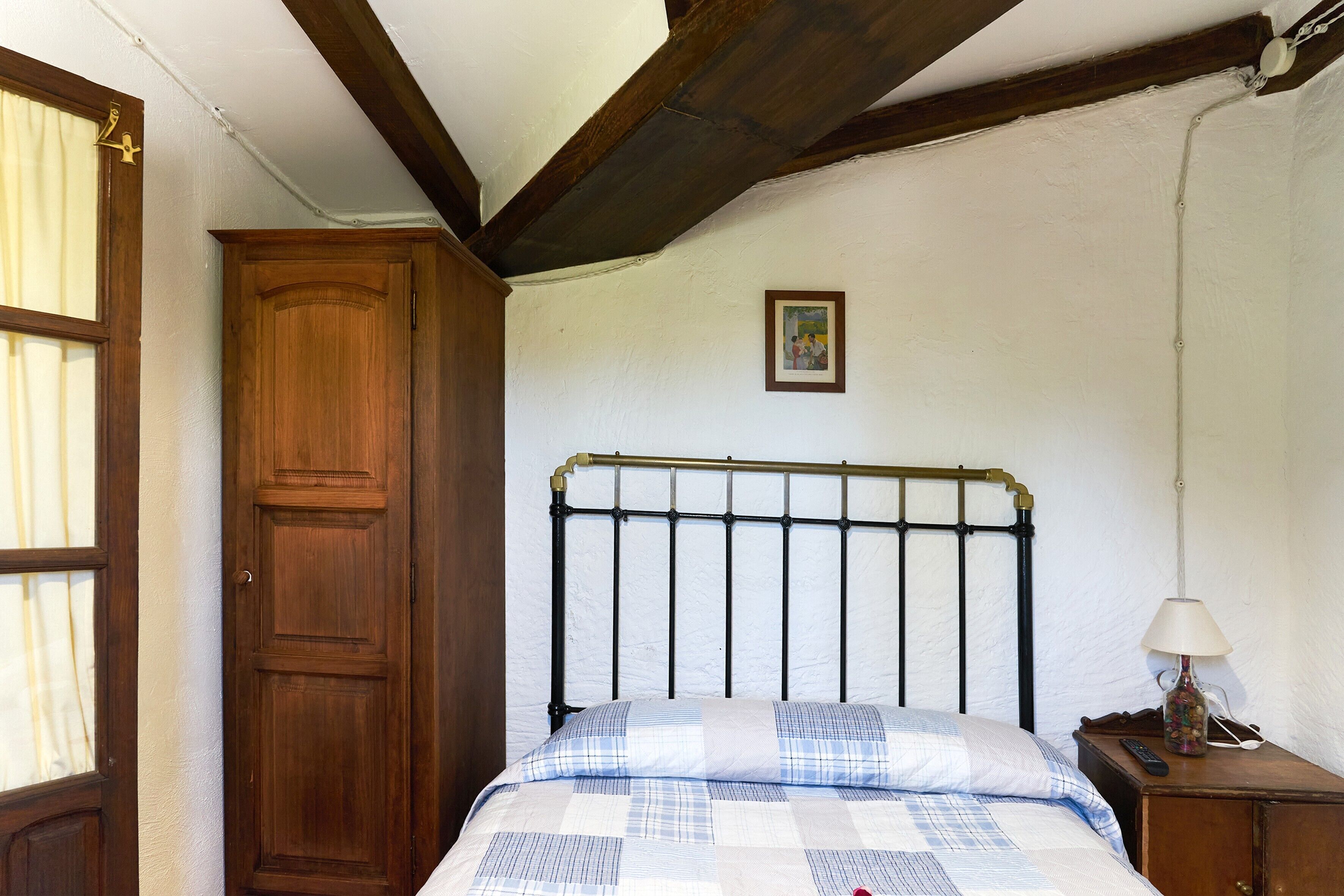 Un dormitorio con puerta de madera, estructura de cama metálica, colcha acolchada, mesita de noche con lámpara y un cuadro enmarcado en la pared.