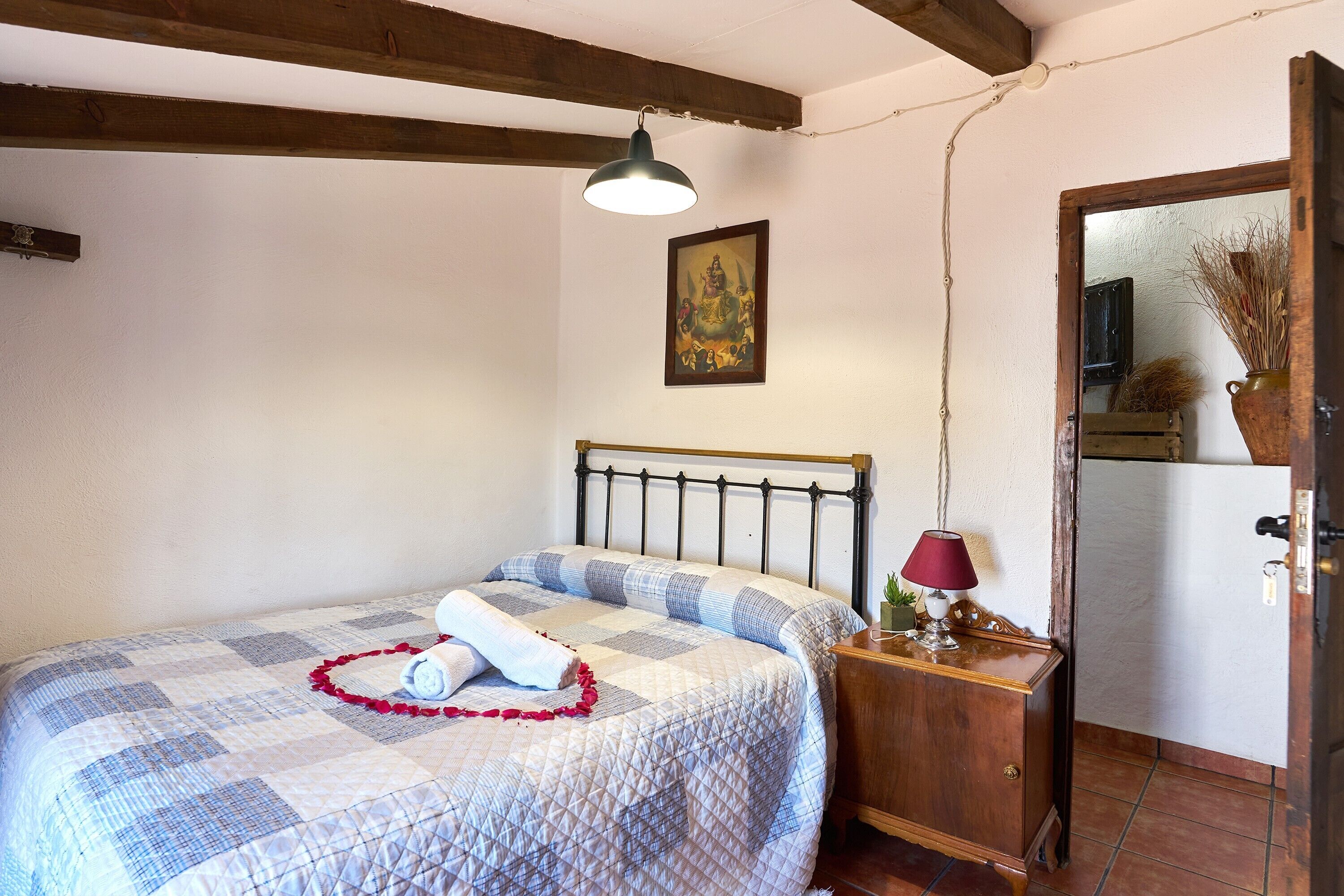 A bedroom with a bed, a wooden nightstand, a lamp, a framed picture on the wall, and a door leading to another room.